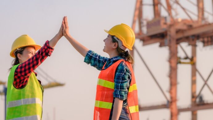 Two female port workers high fiving at a port with a crane in the background
