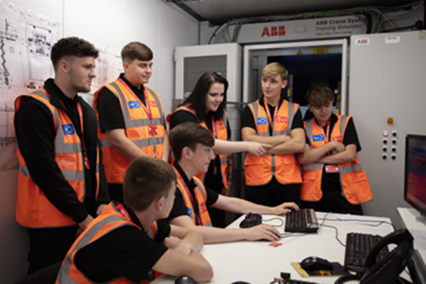 A group of apprentices in hi-vis vests are around a desk looking at a monitor