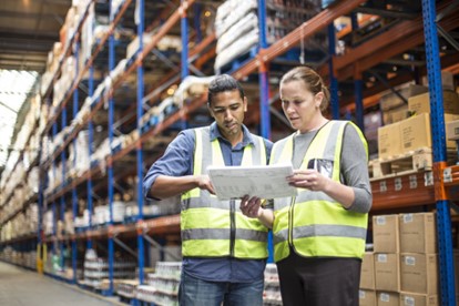 Two workers in a warehouse looking over paperwork and wearing hi vis vests